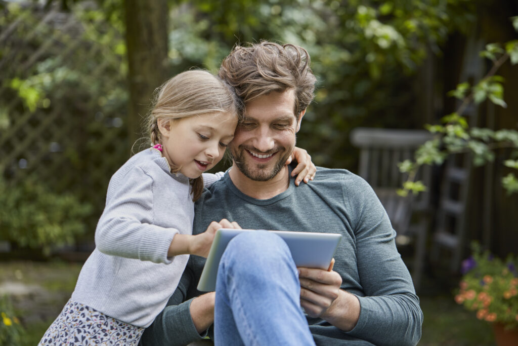Ein fröhlicher Vater und seine kleine Tochter sitzen mit einem Tablet im Garten