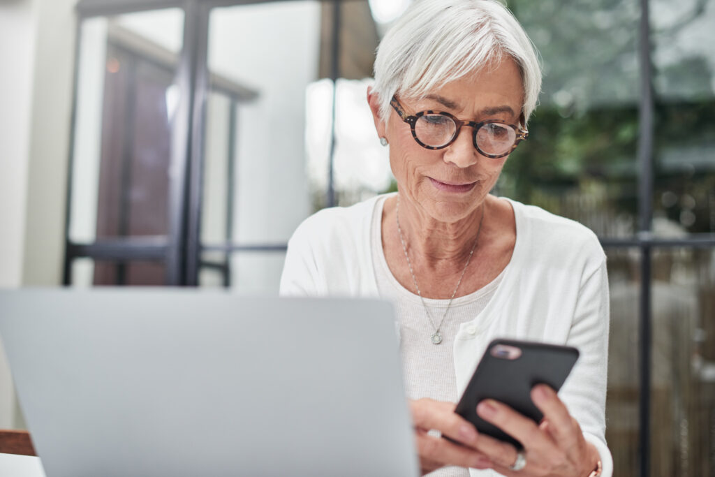 Eine ältere Frau am Laptop blickt skeptisch auf ihr Smartphone