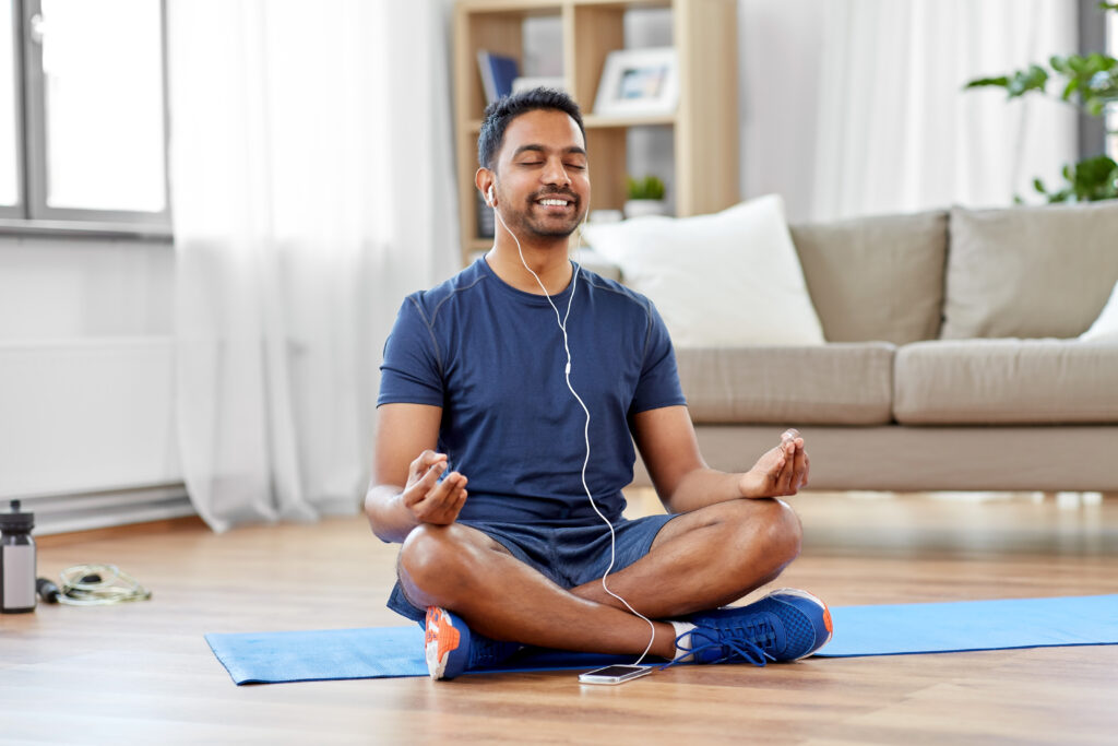 Junger Mann sitzt mit geschlossenen Augen und Kopfhörern auf einer Yogamatte im Wohnzimmer und meditiert.